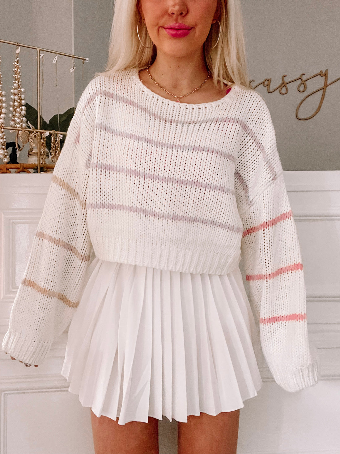 Cream-colored knit striped sweater with pink and beige accents, paired with a white pleated skirt.
