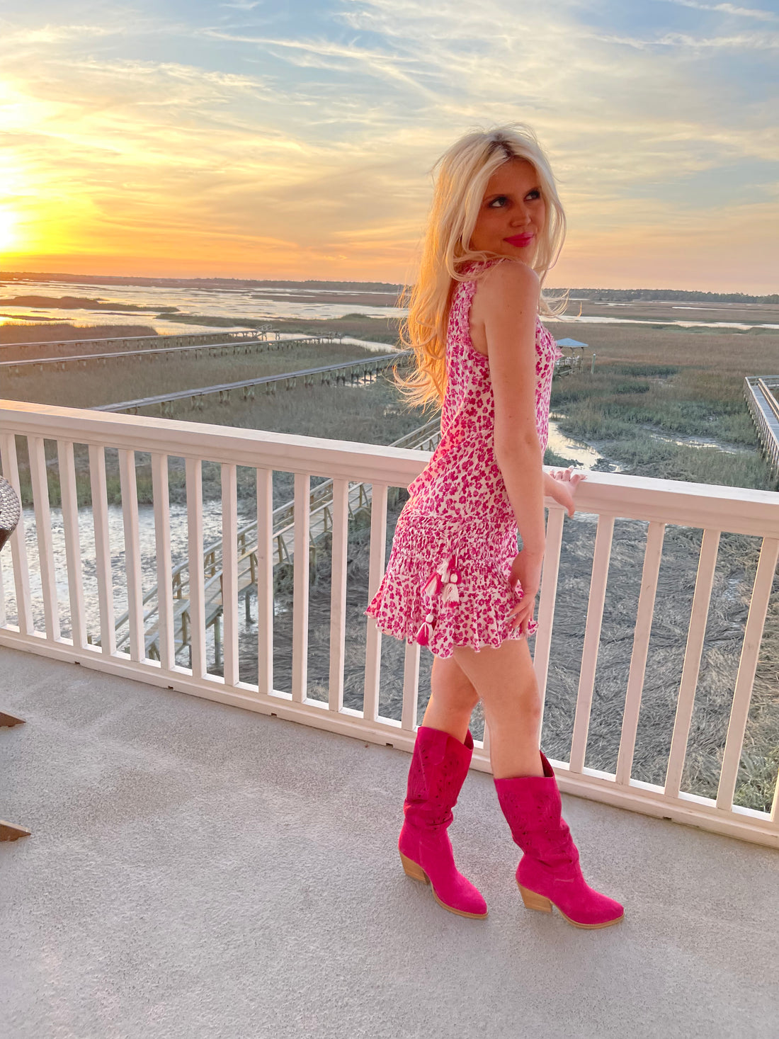 Hot pink suede boots on a deck overlooking a sunset marsh vista.
