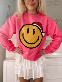 Hot pink crewneck sweatshirt featuring a cheerful yellow smiley face wearing a Santa hat.
