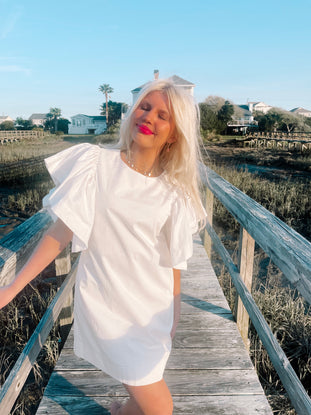 White puff-sleeve mini dress on a wooden boardwalk, sunny coastal background.
