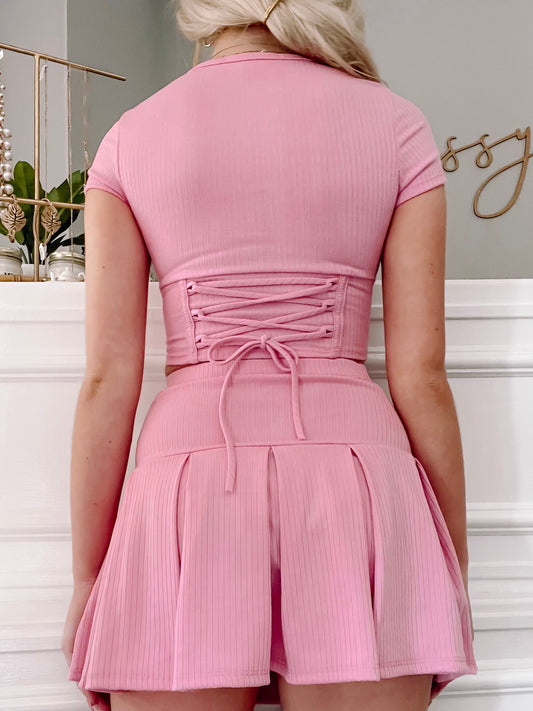 Pink ribbed knit crop top and pleated mini skirt set; back view showcasing lace-up detail.
