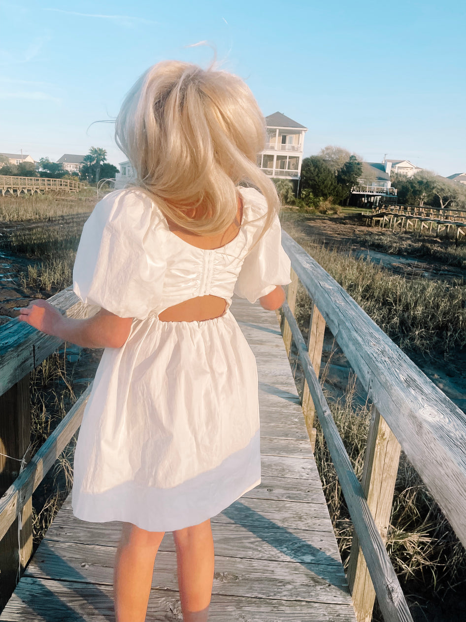 Off-white puff-sleeve mini dress with open back detail, worn outdoors on a wooden boardwalk.
