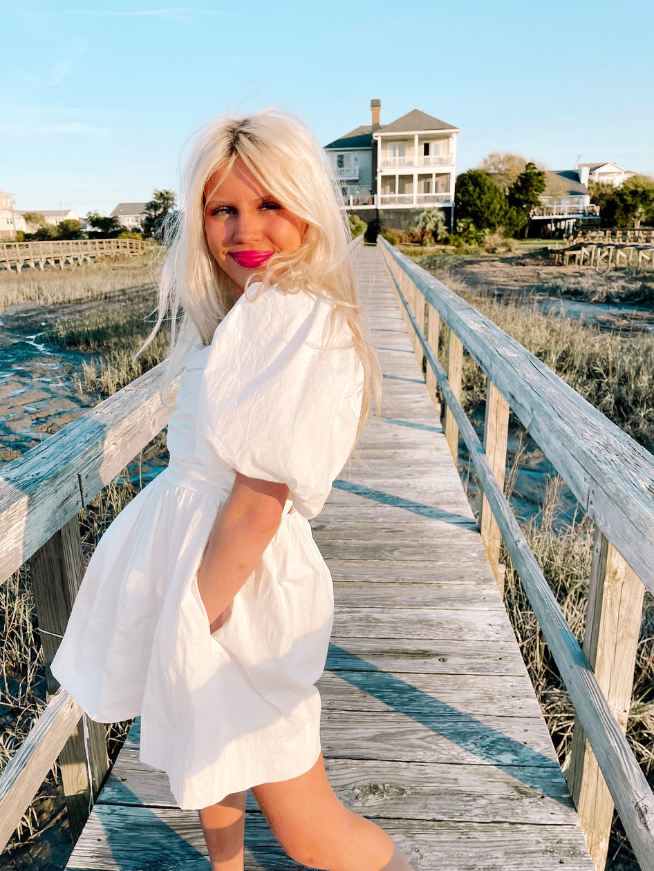 Woman in white puff-sleeve mini dress on a boardwalk overlooking coastal marsh.
