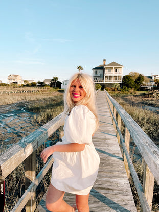 Woman in white puff-sleeve mini dress on a coastal boardwalk.
