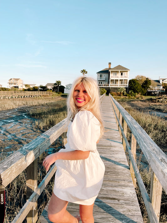 Woman in white puff-sleeve mini dress on a coastal boardwalk.