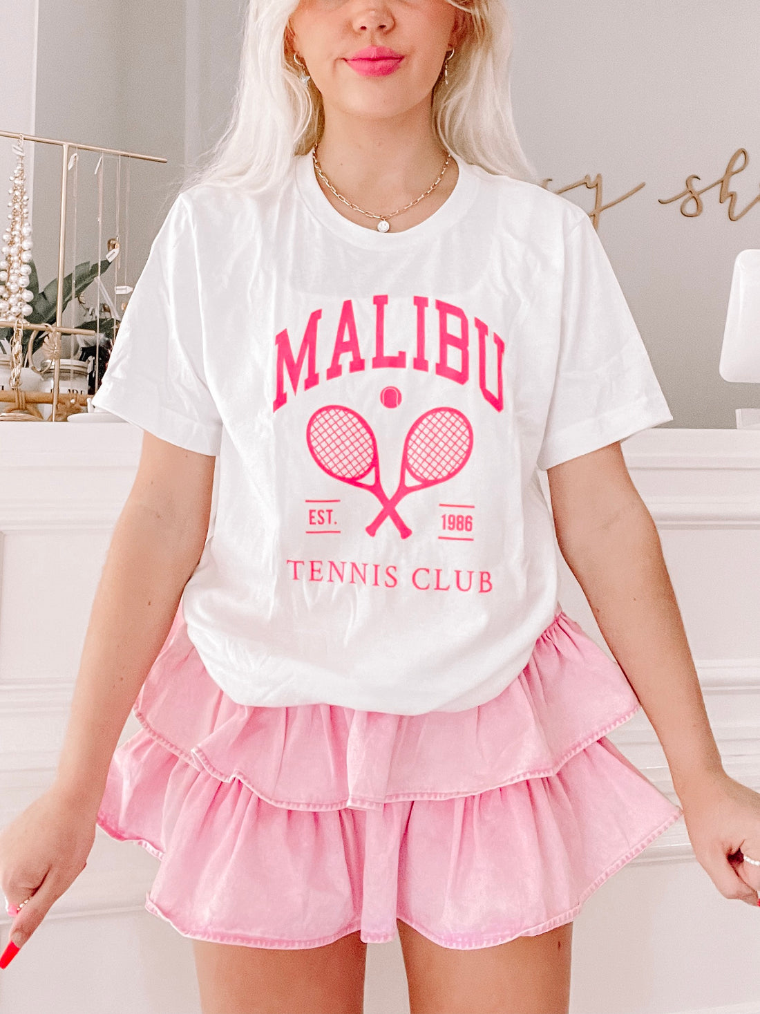 White Malibu Tennis Club tee with pink logo, tennis rackets, and "Est. 1986" detail.
