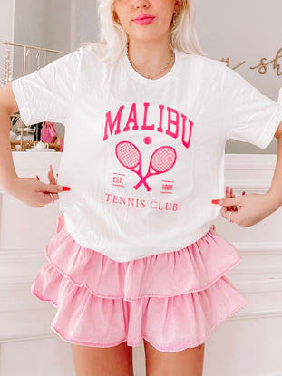 White tee featuring pink tennis-themed graphic, modeled with pink ruffle shorts.
