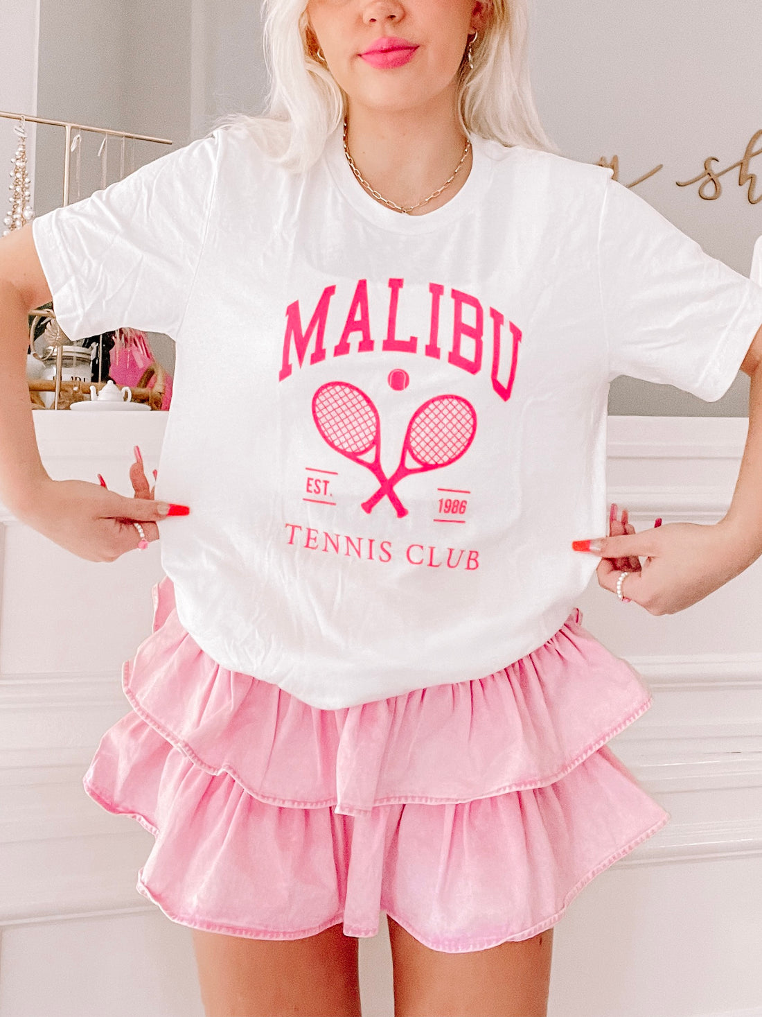 White tee featuring pink tennis-themed graphic, modeled with pink ruffle shorts.
