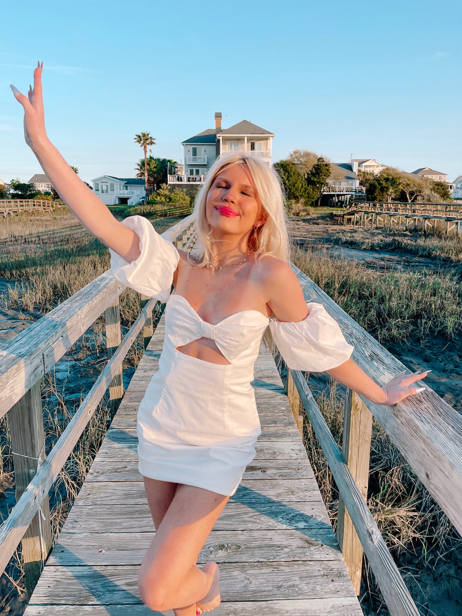 Woman in white cutout dress on a boardwalk overlooking the water.
