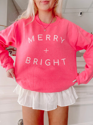 Hot pink crewneck sweatshirt with “Merry + Bright” in white lettering, styled with a white ruffle skirt.
