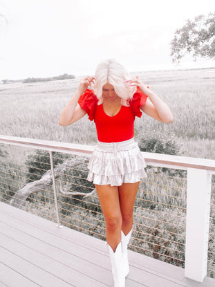 Red ruffled bodysuit styled outdoors, paired with a metallic skirt and white boots.
