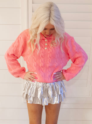 Pink cable-knit sweater styled with a shimmering silver mini skirt against a white wood backdrop.
