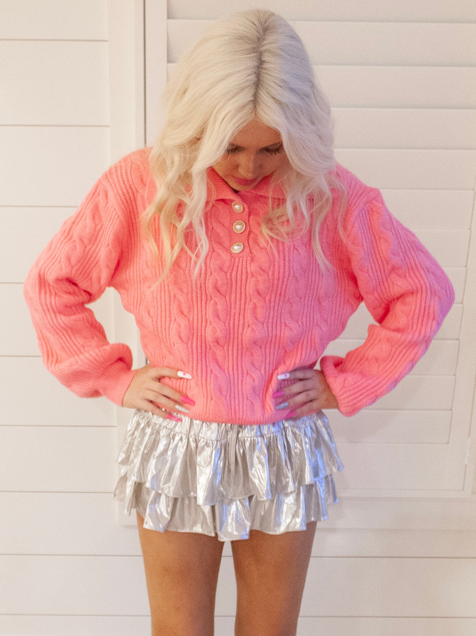 Pink cable-knit sweater styled with a shimmering silver mini skirt against a white wood backdrop.
