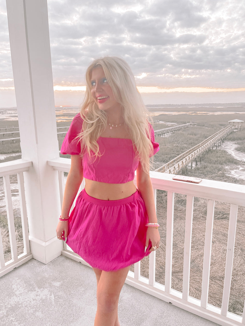 Hot pink mini skirt and matching crop top worn outdoors on a porch overlooking marshland at sunset.
