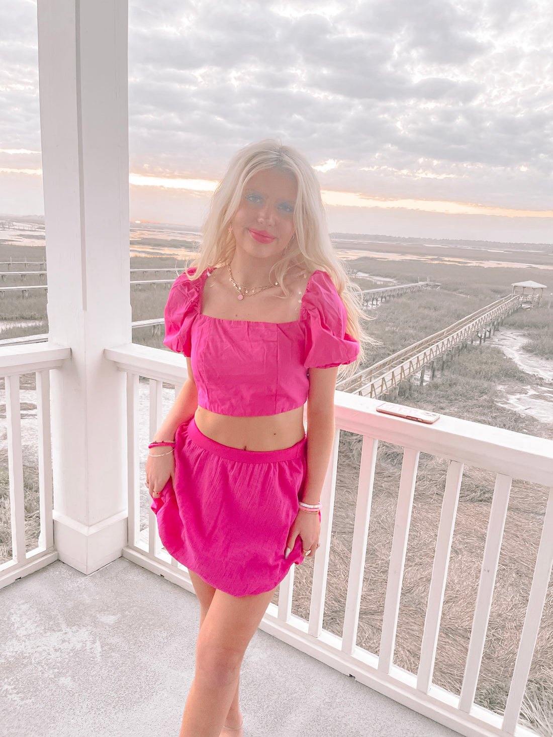 Hot pink mini skirt and matching crop top worn outdoors on a porch overlooking a marsh at sunset.
