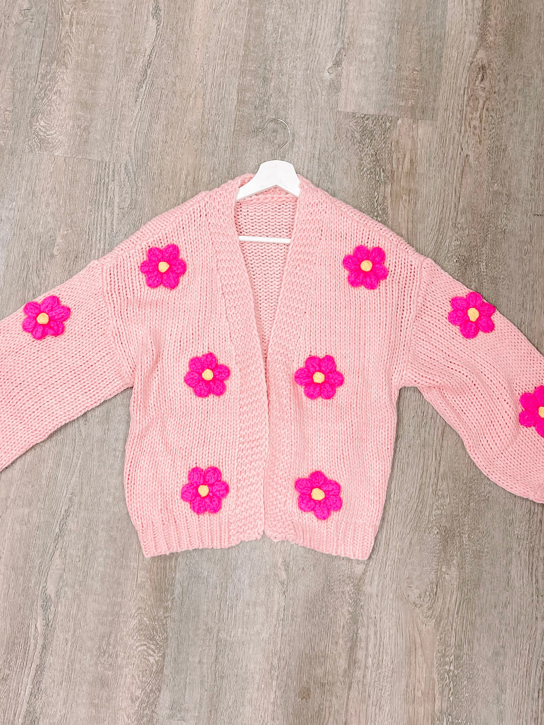 Pink knit cardigan adorned with bright pink flowers, hanging on a white hanger against a wood background.
