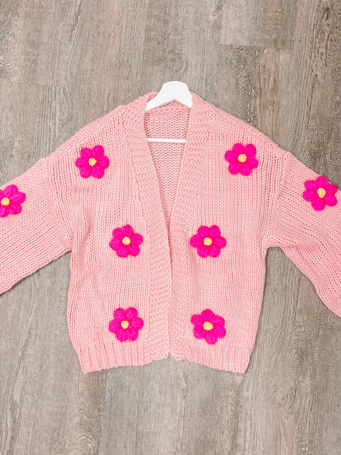 Pink knit cardigan adorned with bright pink flower appliqués, displayed on a hanger.
