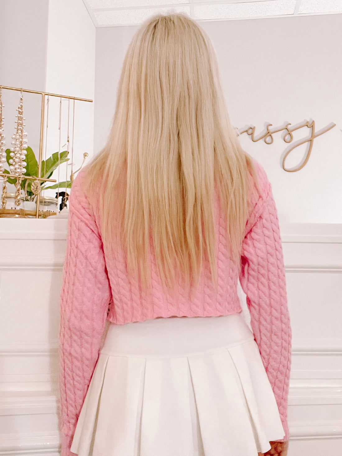 Pink cable-knit cropped sweater shown from the back, paired with a white pleated skirt.

