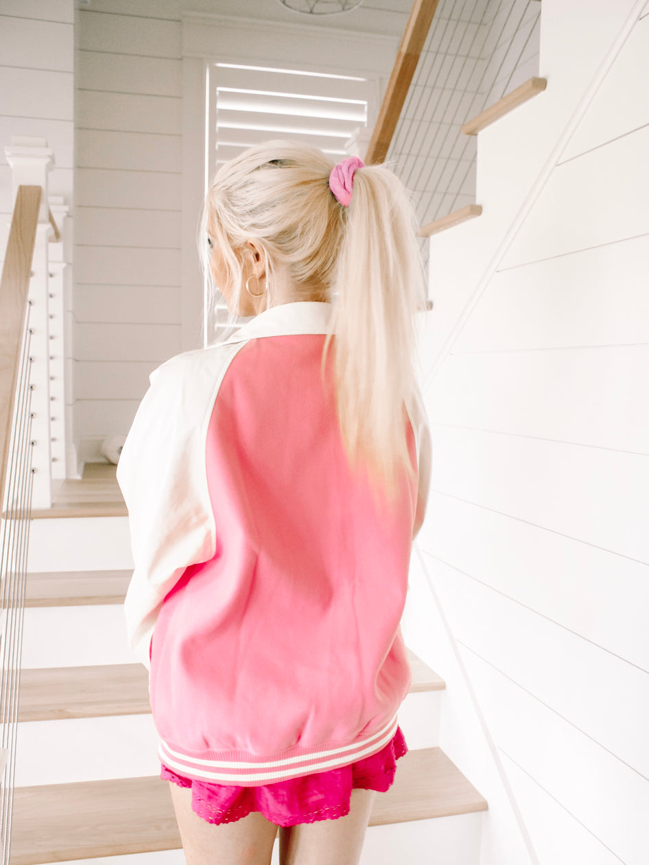 Pink and white varsity-style jacket shown from the back, worn indoors on a staircase.
