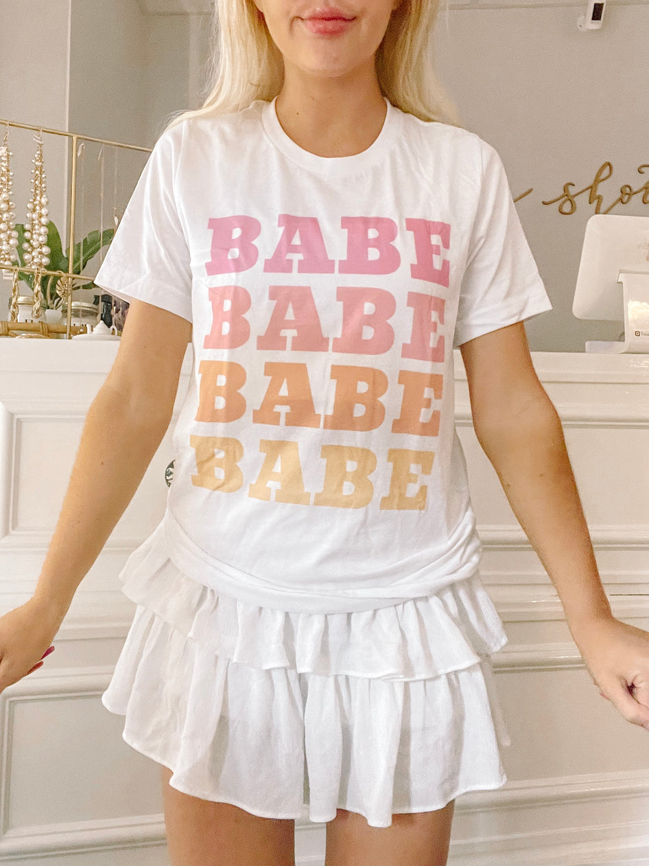 White cotton tee featuring a stacked "Babe" graphic in pastel shades, modeled with a white ruffle skirt.
