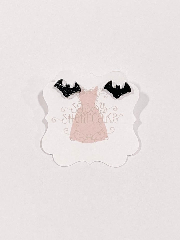 Black glitter bat stud earrings displayed on a pink card
