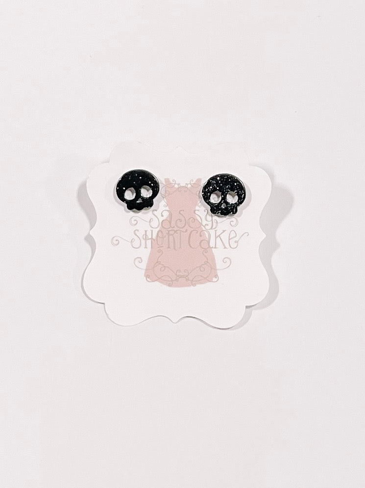 Black glitter skull stud earrings displayed on a pink tag.
