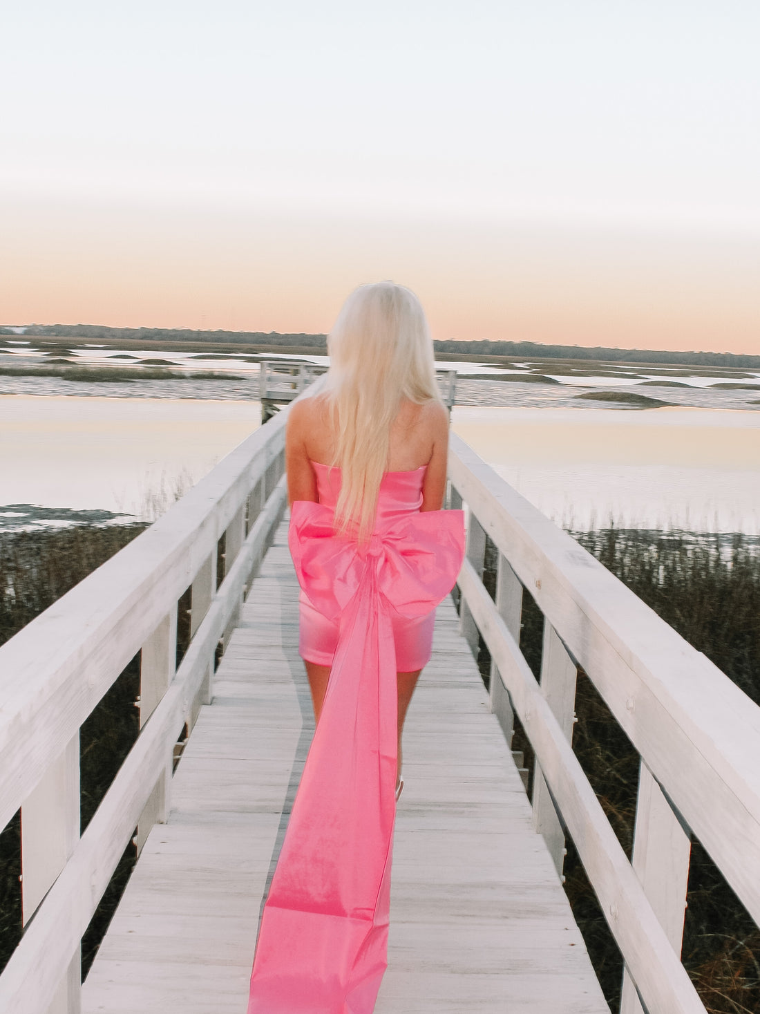 Pink mini dress with large bow detail, trailing along a boardwalk overlooking tranquil water at sunset.
