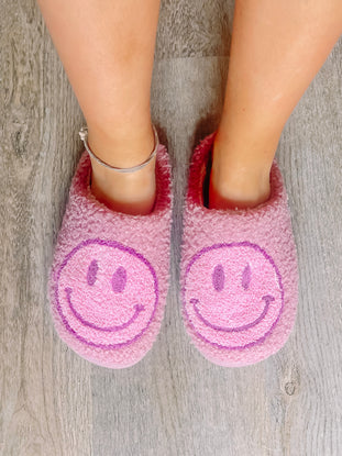 Pink fuzzy Sassy Slumber slippers with purple smiley faces, perfect for cozy nights.
