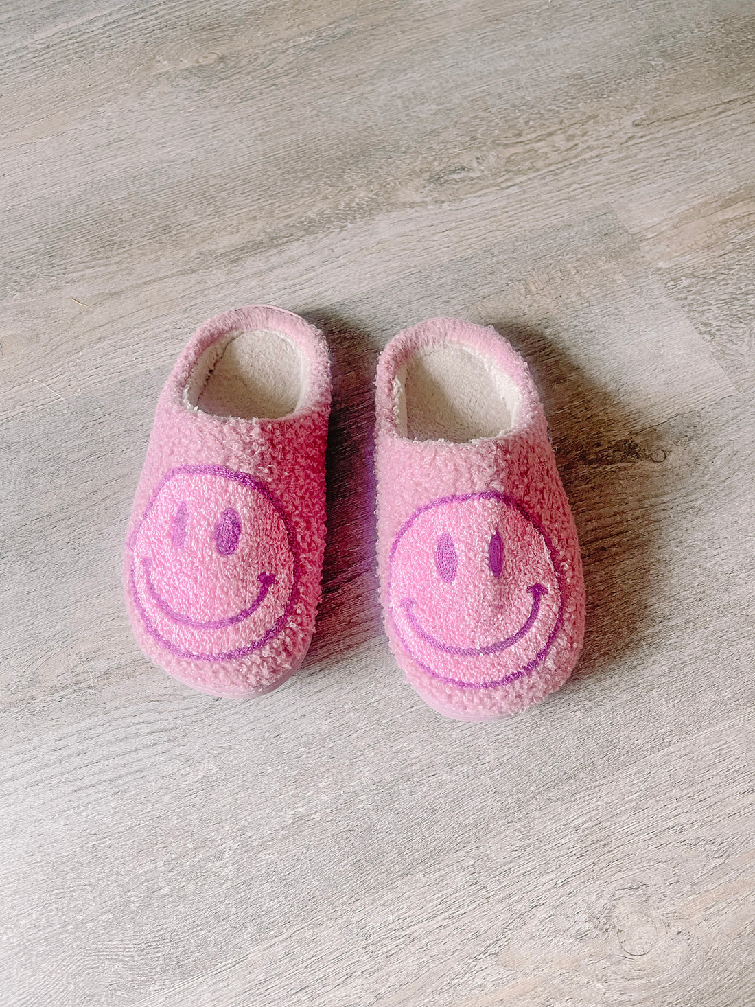 Pink fuzzy slippers with purple smiley faces, on a wood floor.
