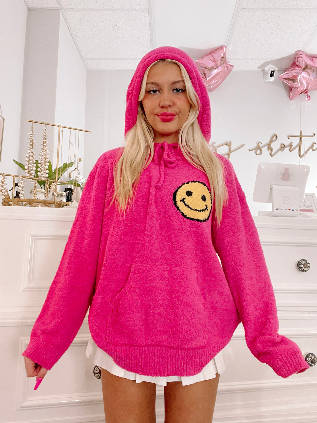 Hot pink fuzzy hoodie sweater featuring a smiling face, modeled indoors.
