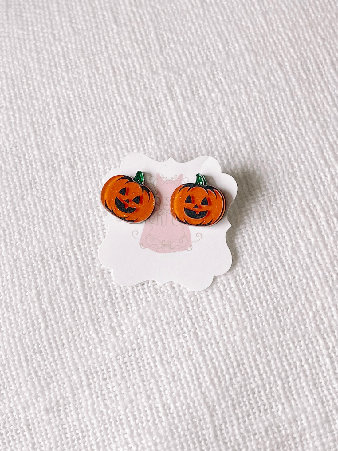 Jack-o'-lantern stud earrings displayed on a card against a textured white background.
