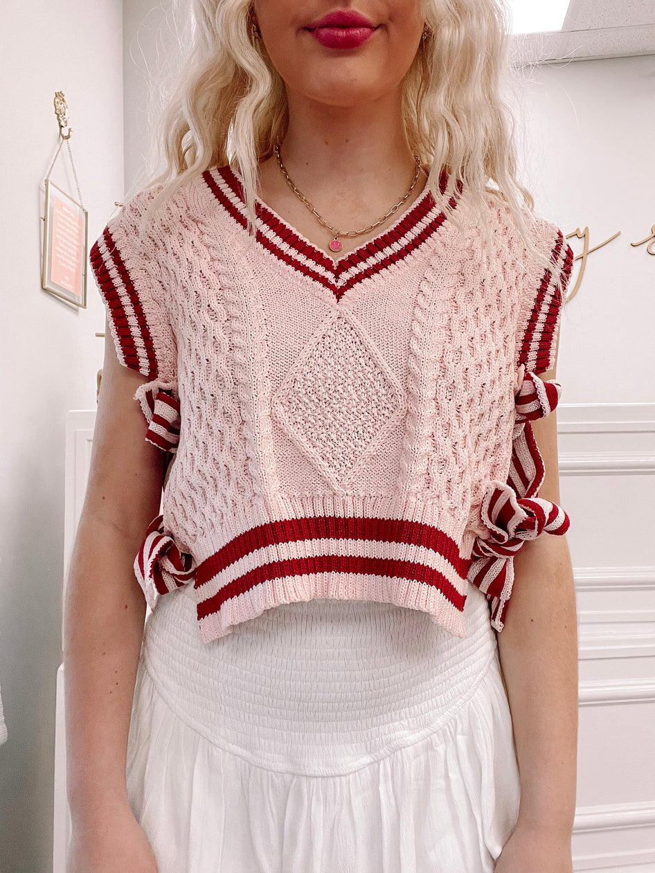 Pink cable knit cropped sweater vest with burgundy stripes, side ties, and a V-neck.
