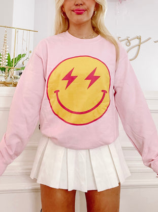 Pink crewneck sweatshirt featuring a yellow smiley face with red lightning bolt eyes.
