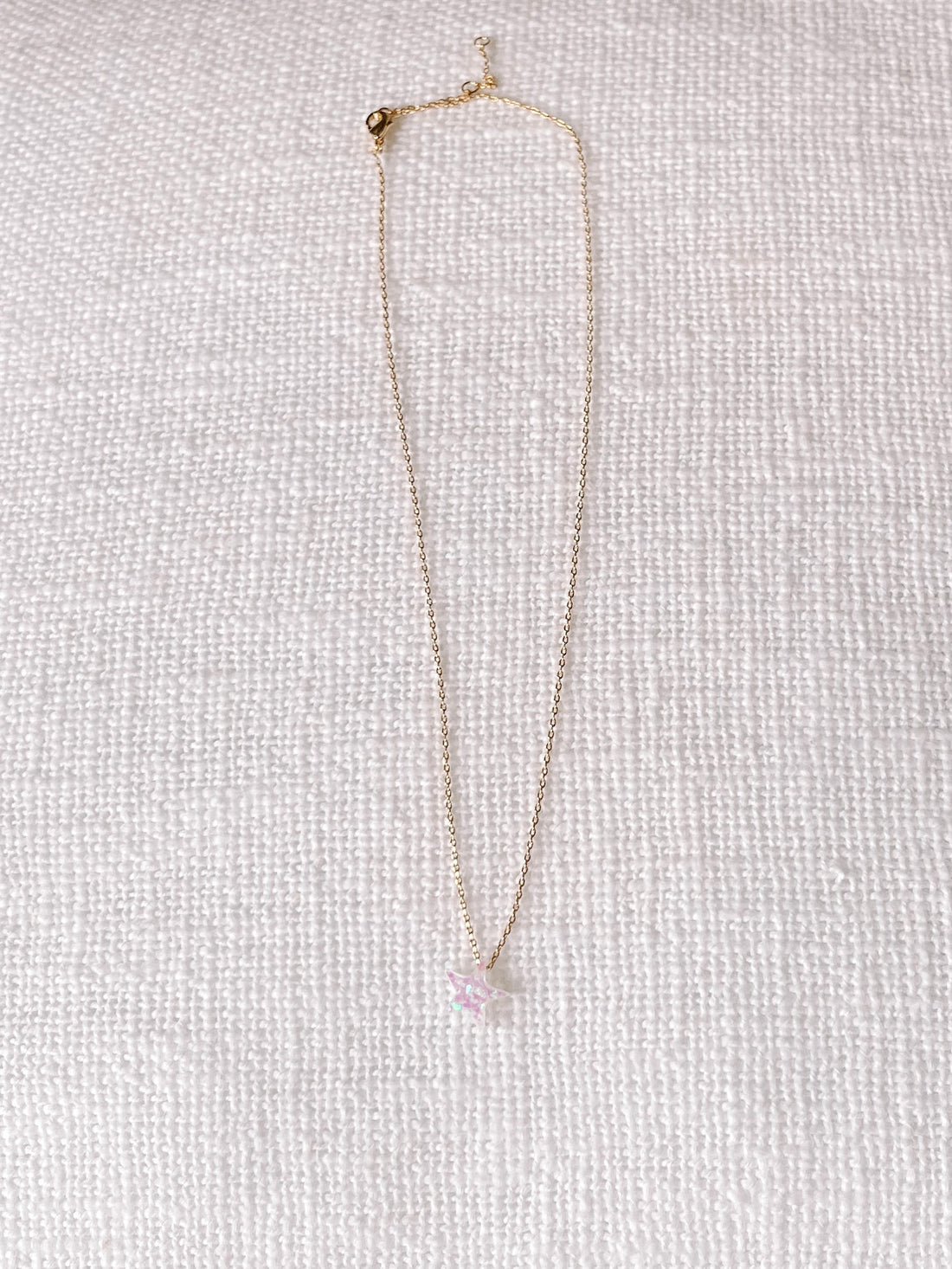 Gold chain necklace featuring a sparkly, pastel pink star charm, displayed on a white textured background.
