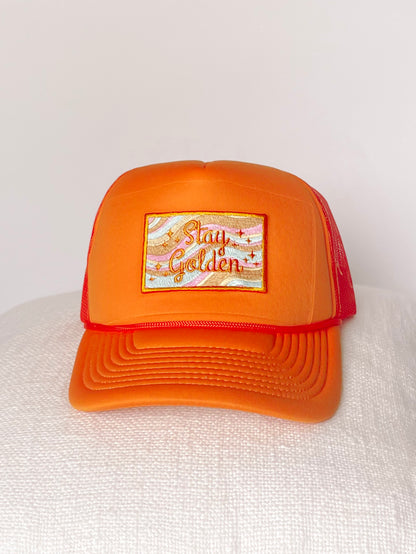 Orange trucker hat with embroidered "Stay Golden" patch; pastel wave design.
