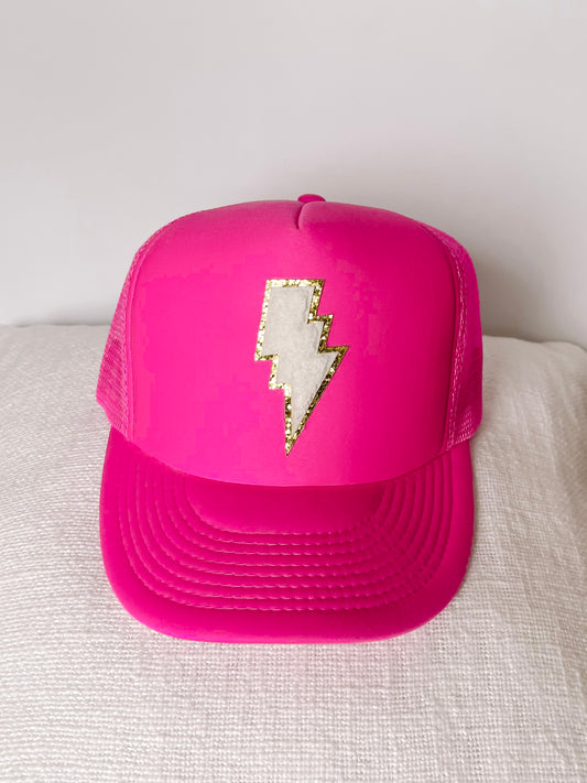 Hot pink trucker hat with a white and gold lightning bolt design, displayed on a white surface.
