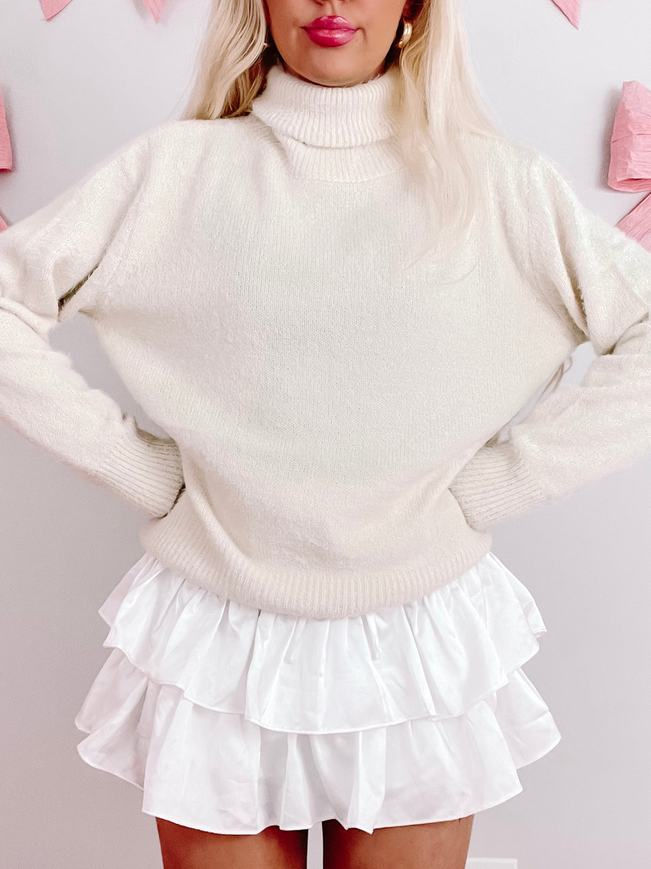 Cream turtleneck sweater styled with a white ruffled mini skirt.
