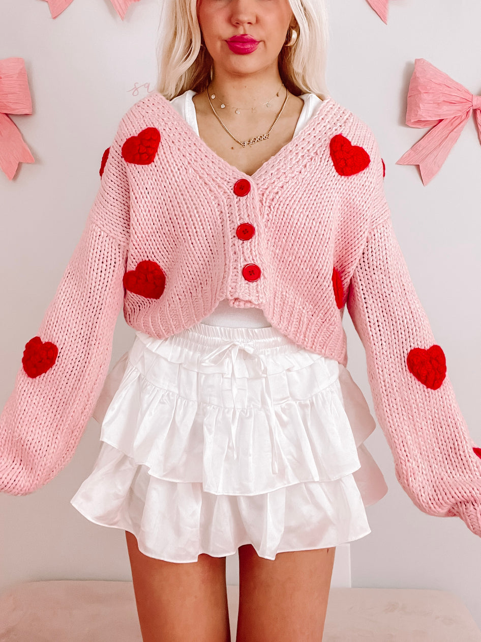 Pink knitted cardigan with 3D red hearts, cropped length, perfect for Valentine's Day.
