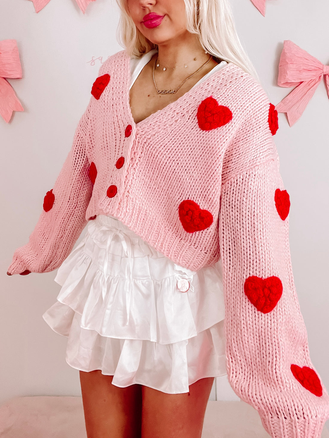 Pink knit cardigan adorned with red felt hearts, modeled with a white ruffled skirt.
