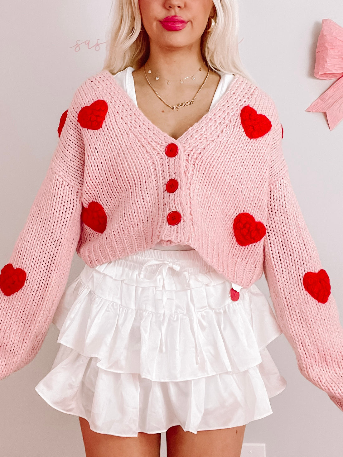 Pink knit cardigan adorned with red heart appliqués, button front, modeled with white tiered skirt.
