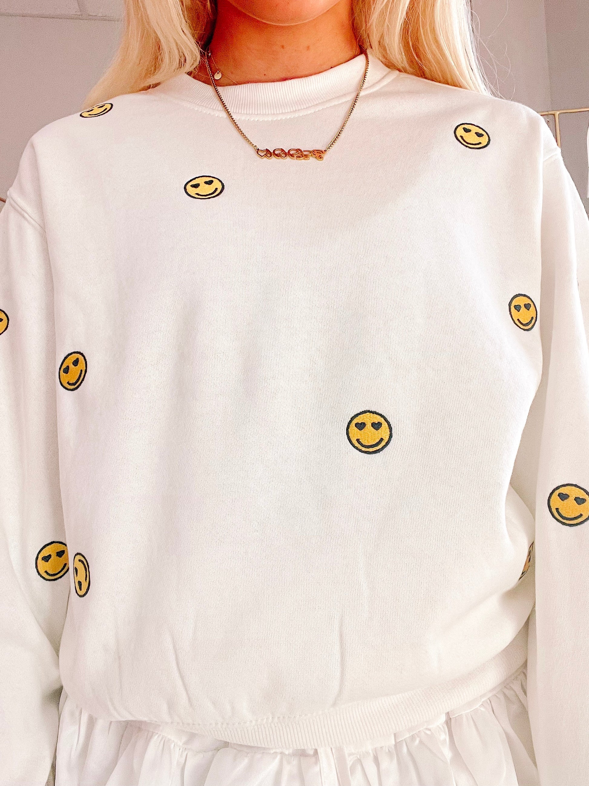 Cream-colored crewneck sweatshirt with embroidered smiley faces;  closeup shows soft fabric texture.
