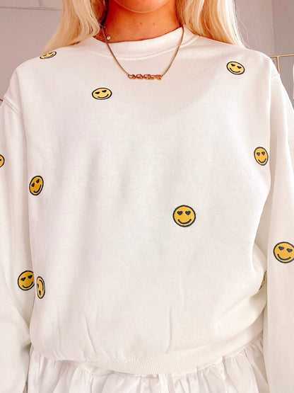 Cream-colored crewneck sweatshirt with embroidered smiley faces;  closeup shows soft fabric texture.
