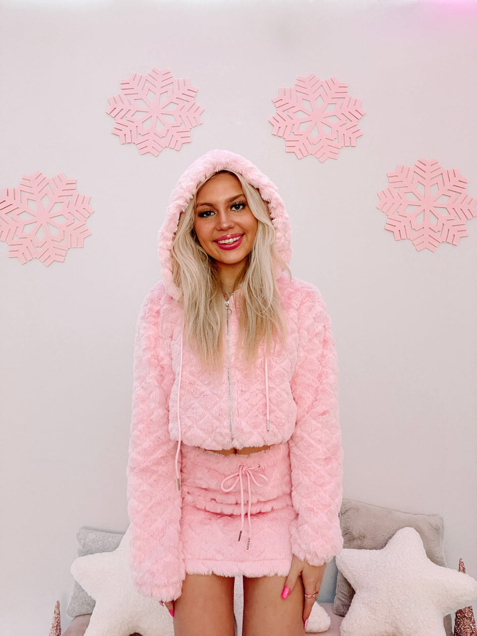 Pink fuzzy hooded zip-up jacket and mini skirt set, modeled against a pastel background.
