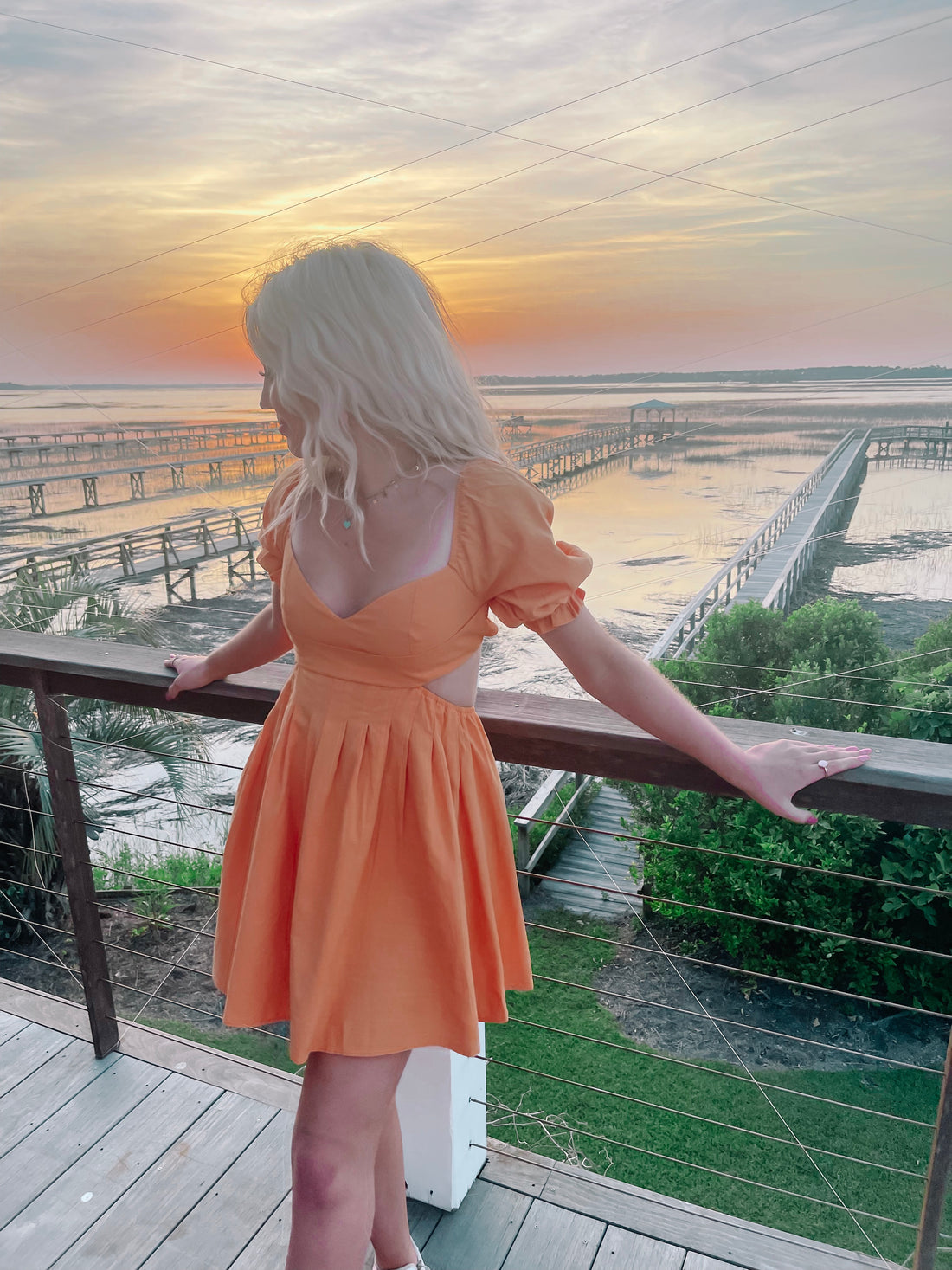Orange sundress worn on a deck overlooking a sunset over calm water and wooden piers.

