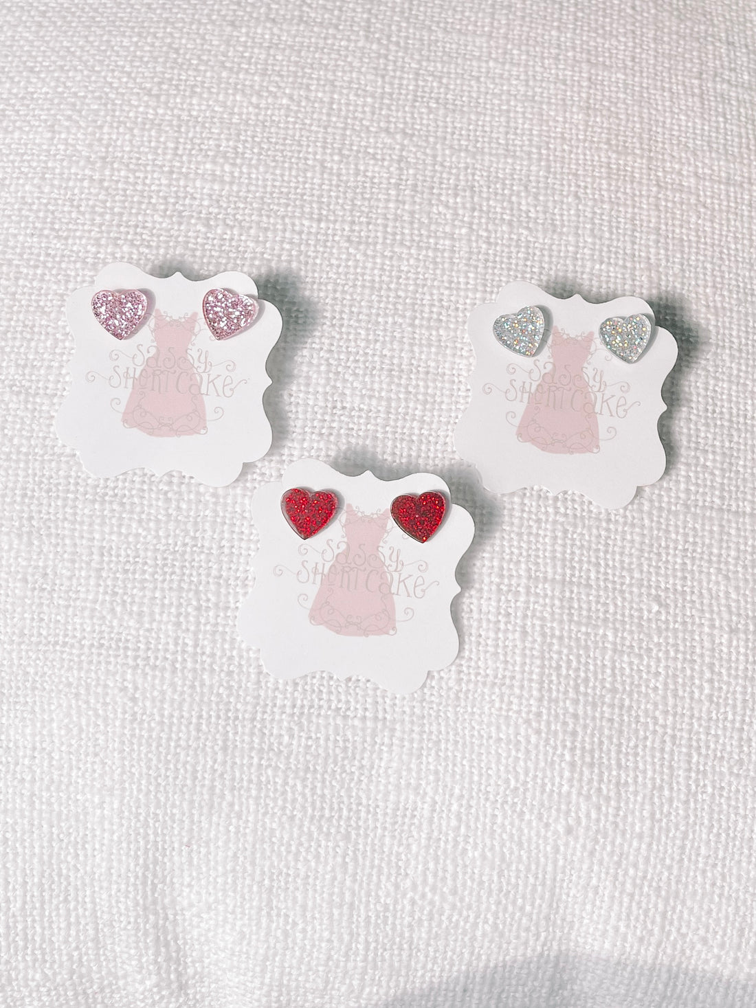 Glittery heart-shaped earrings displayed on individual cards, pink, silver, and red.

