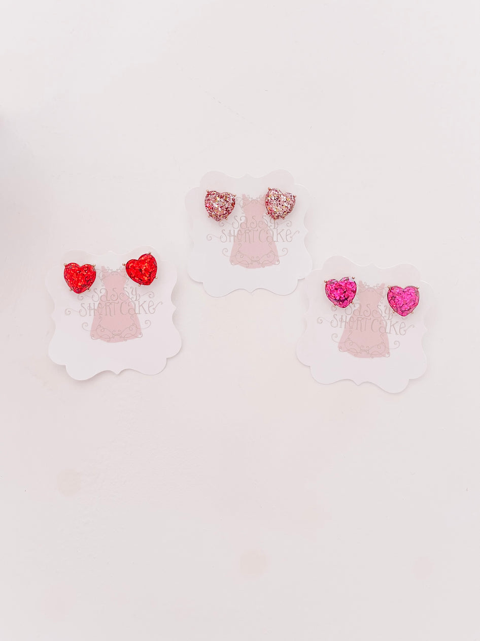 Sparkly heart-shaped earrings in red, pink, and rose gold displayed on individual cards.

