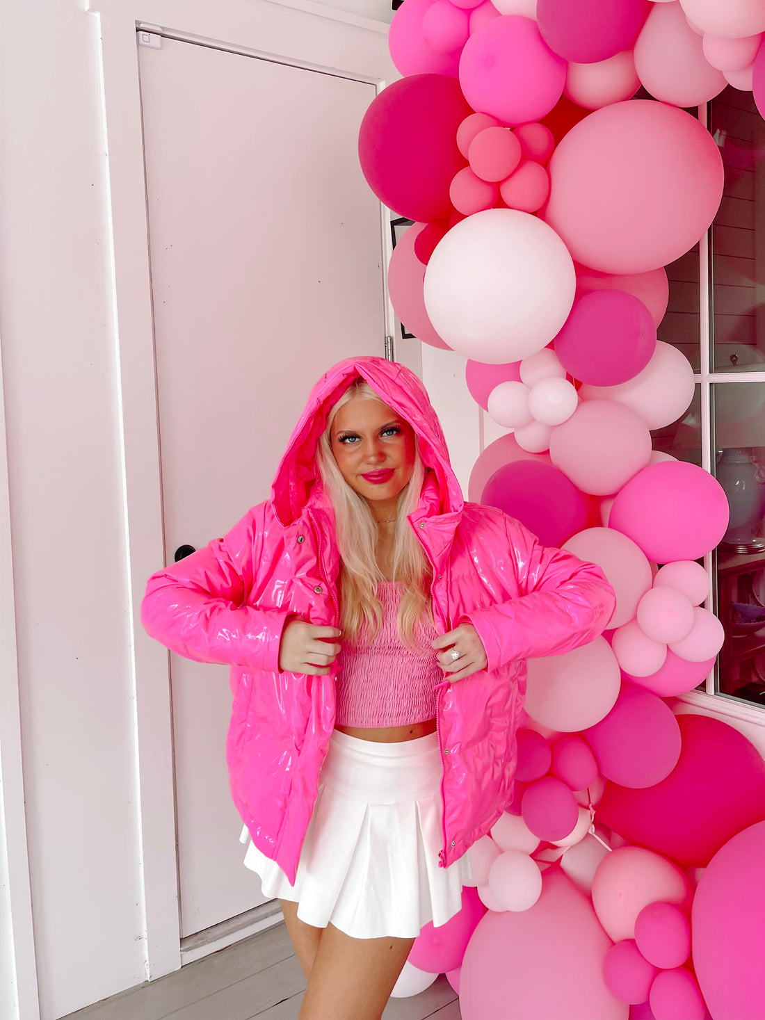 Bright pink shiny puffer jacket with a hood, modeled against a pink balloon backdrop.
