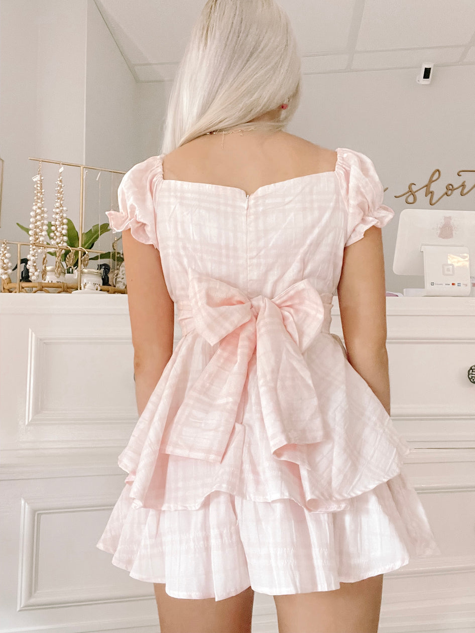 Pink gingham tiered romper with puff sleeves and a large bow at the waist, shown from the back.
