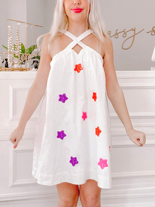 White eyelet sleeveless dress adorned with colorful star embellishments, shown on a model against a white wall.
