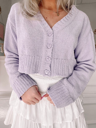 Lavender plush cropped cardigan with heart-shaped buttons, modeled over a white tiered skirt.
